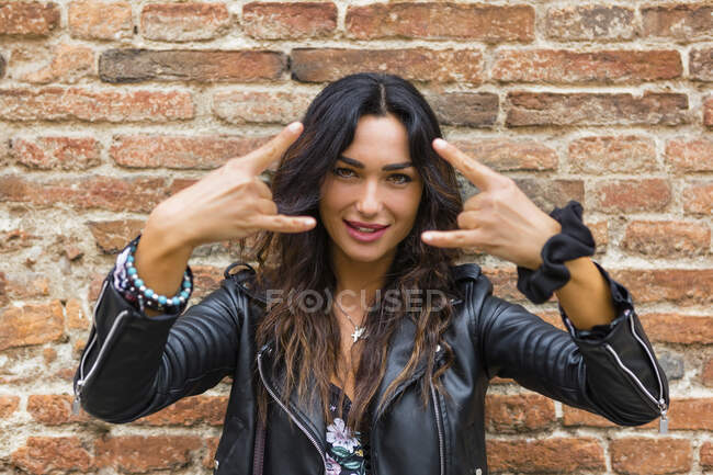 Retrato de jovem mulher vestindo jaqueta de couro preto e mostrando sinal de rock and roll, parede de tijolo — Fotografia de Stock
