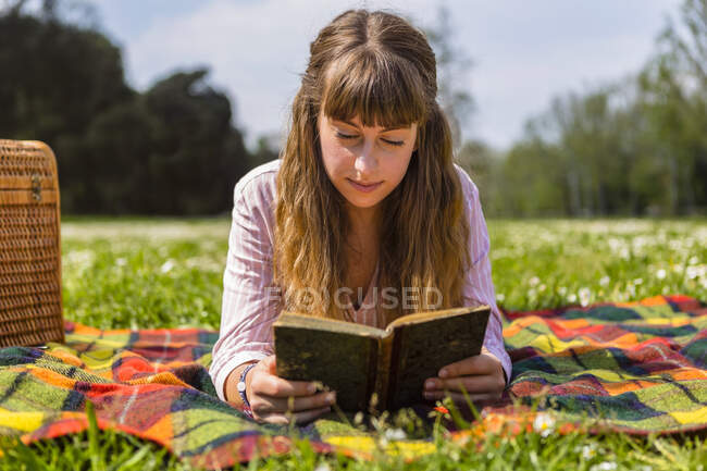 Молода жінка лежить на пікніковому покритті і читає книжку в парку. — стокове фото