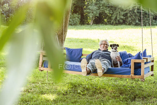 Homem sênior relaxando em uma cama de balanço em seu jardim com seu cão — Fotografia de Stock