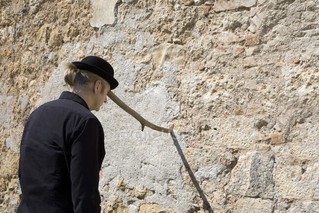 Man wearing a bowler hat balancing a stick at a stone wall — Stock Photo