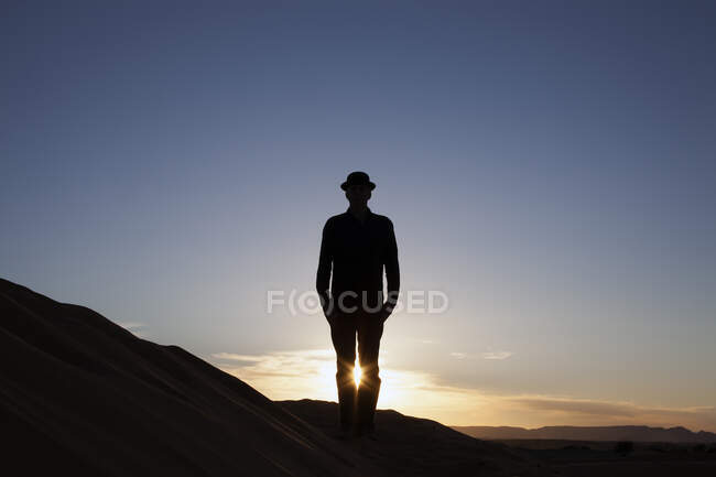 Marruecos, Merzouga, Erg Chebbi, silueta de hombre con sombrero de jugador de bolos parado en la duna del desierto al atardecer - foto de stock