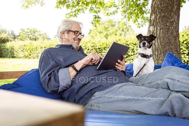 Homem sênior relaxando em uma cama de balanço em seu jardim, usando tablet digital — Fotografia de Stock