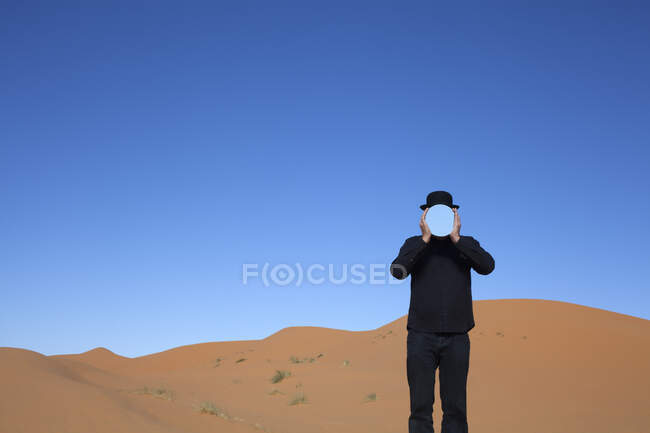Morocco, Merzouga, Erg Chebbi, man wearing a bowler hat holding mirror in front of his face in desert dune — Stock Photo