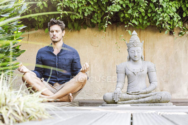 Hombre sentado con las piernas cruzadas junto a la estatua de Buda en un jardín zen, meditando - foto de stock