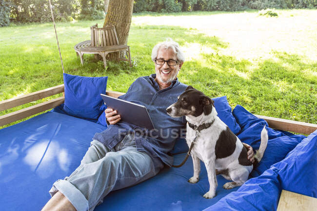 Homem sênior relaxando em uma cama de balanço em seu jardim, usando tablet digital — Fotografia de Stock