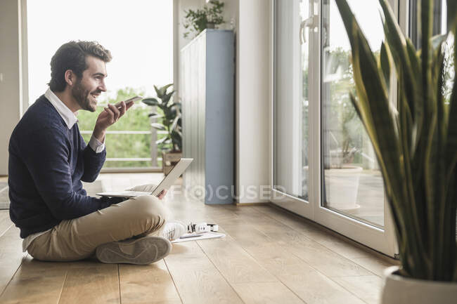 Young man sitting cross-legged in front of window, using laptop, talking on the phone — Stock Photo