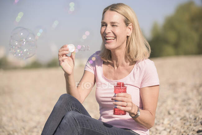 Glücklich reife Frau pustet Seifenblasen auf Kieselstrand — Stockfoto