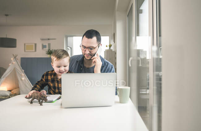 Padre trabajando en la mesa en la oficina del hogar con su hijo sentado en su regazo - foto de stock