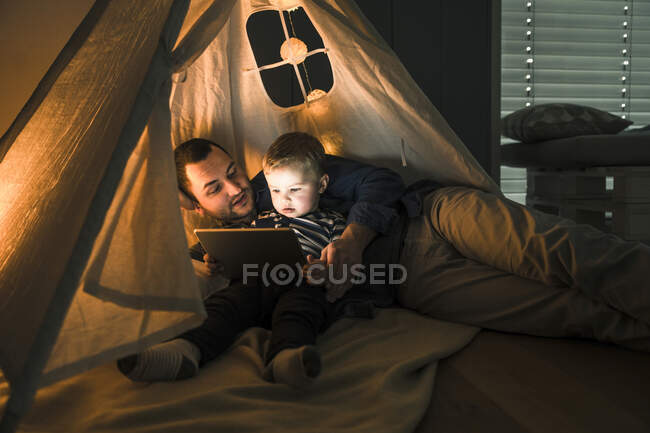 Pai e filho compartilhando um tablet em uma tenda escura em casa — Fotografia de Stock
