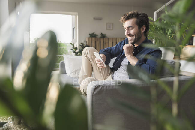 Giovane seduto sul divano di casa, utilizzando lo smartphone — Foto stock