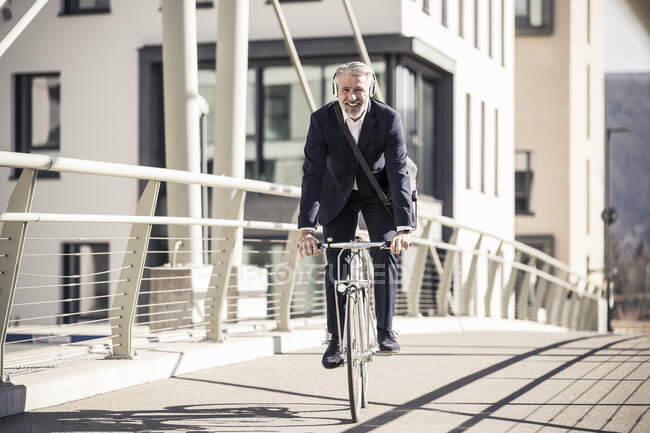 Hombre de negocios maduro sonriente con auriculares montando bicicleta en un puente en la ciudad - foto de stock