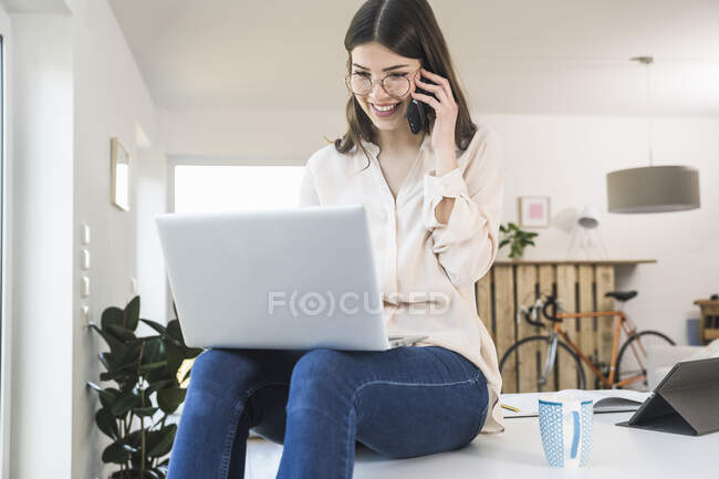 Усміхаючись, молода жінка сидить вдома за столом за допомогою ноутбука і мобільного телефону. — стокове фото