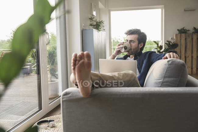 Молодой человек сидит на диване, пользуется ноутбуком, пьет кофе — стоковое фото