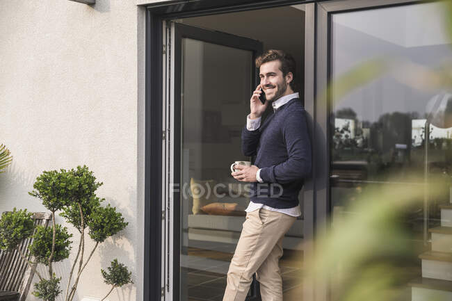 Молодий чоловік нахилився в двері свого дому, тримаючи чашку кави, за допомогою смартфона. — стокове фото