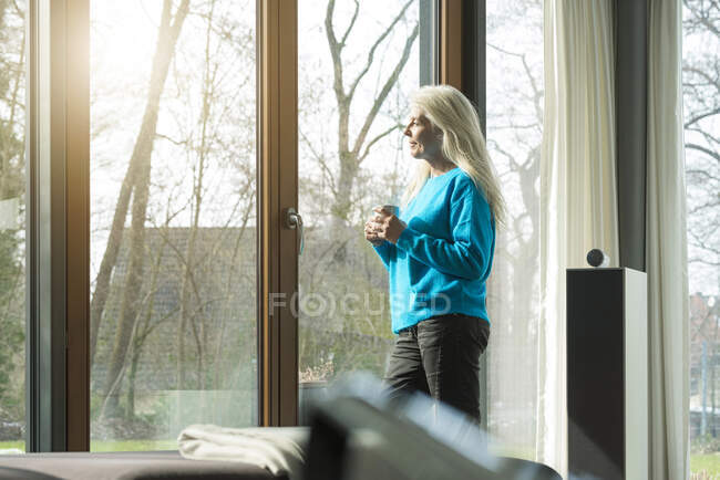 Contenuto donna matura con tazza di caffè appoggiata alla porta della terrazza guardando la distanza — Foto stock