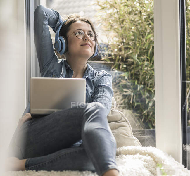 Жінка з навушниками і ноутбуком сидить у вікні вдома. — стокове фото