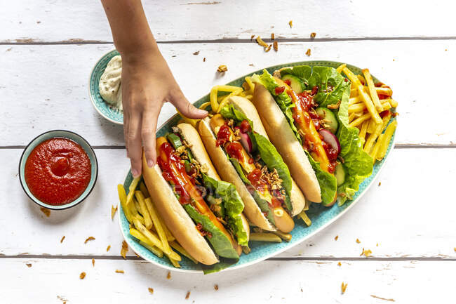 Perros calientes con papas fritas, ketchup y mayonesa, mano tomando un hot dog - foto de stock