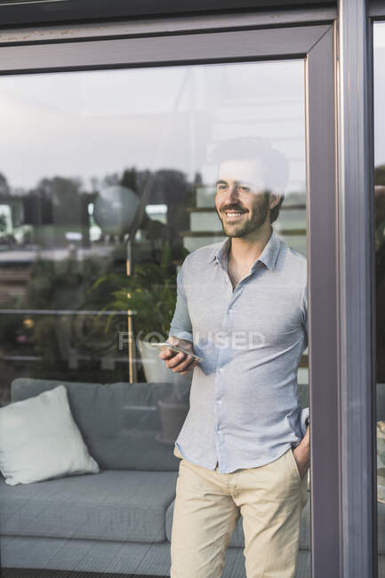 Young man looking out of window, using smartphone — Stock Photo