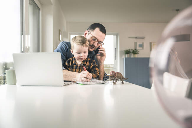 Ocupado pai trabalhando à mesa no escritório em casa com o filho sentado em seu colo — Fotografia de Stock