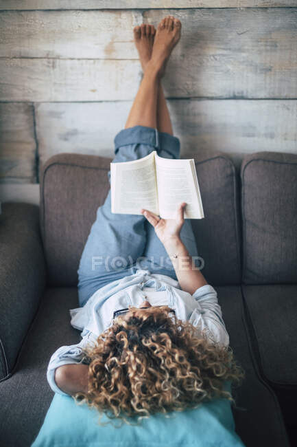 Woman lying on couch at home reading a book — Stock Photo