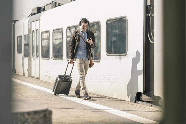 Joven sonriente con auriculares, teléfono celular y maleta caminando en la plataforma de la estación — Stock Photo