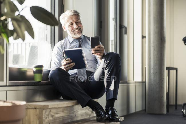 Усміхнений дорослий бізнесмен сидить біля вікна з мобільним телефоном і записником. — стокове фото