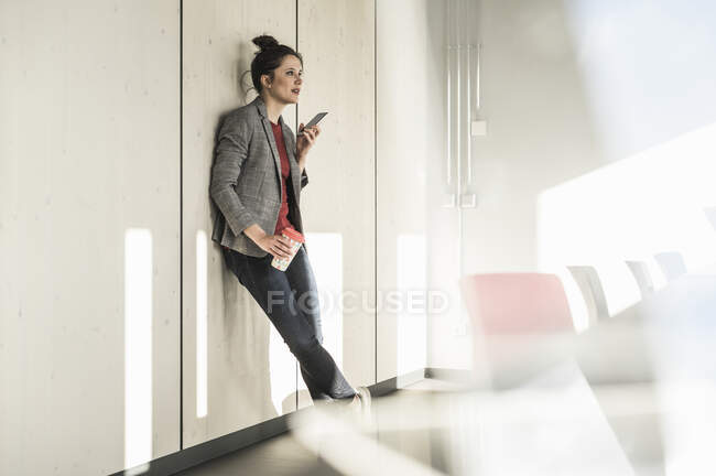 Donna d'affari appoggiata a un muro in ufficio utilizzando il telefono cellulare — Foto stock