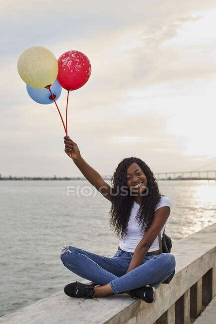 Mocthe, Maputo, retrato de una joven feliz con tres globos sentados frente al mar - foto de stock