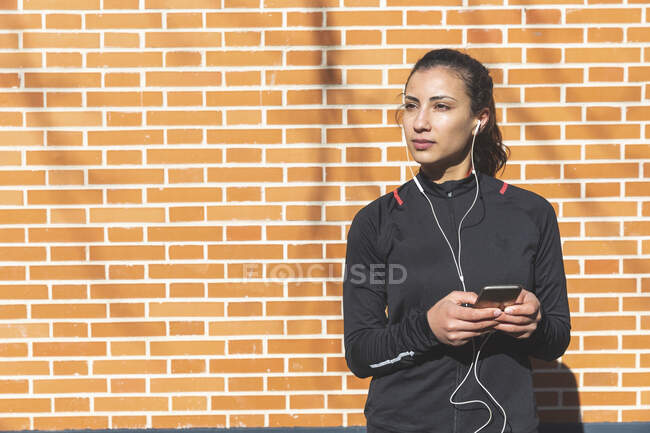 Retrato de mujer joven y deportiva con auriculares y teléfono celular en una pared de ladrillo - foto de stock
