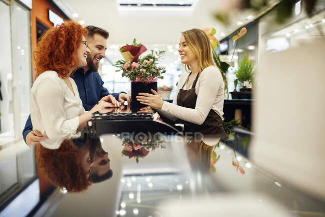 Pareja feliz y asistente de tienda con planta en maceta en el mostrador en la tienda de flores - foto de stock