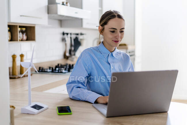 Woman working on laptop at home with wind turbine model on kitchen counter — Stock Photo