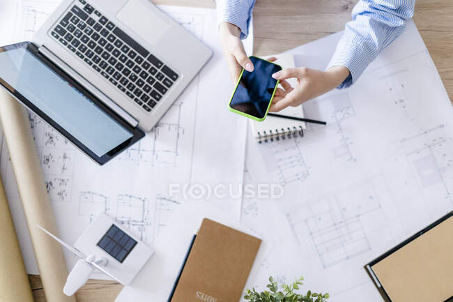 Gros plan de la femme au bureau à l'aide d'un téléphone cellulaire avec plan, ordinateur portable et éolienne sur la table — Photo de stock