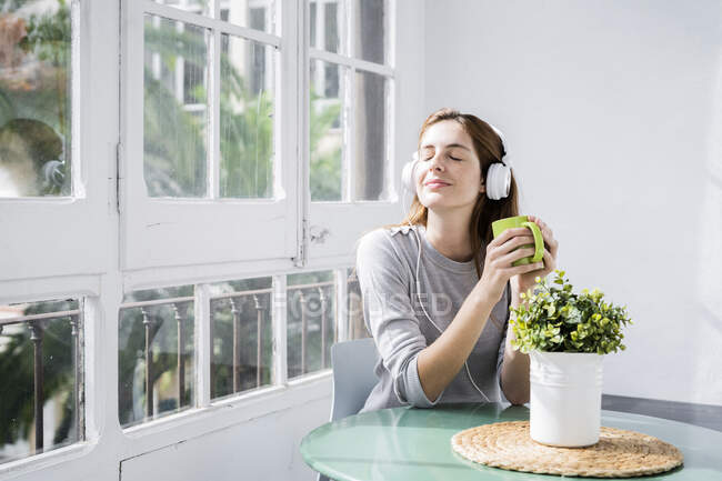 Jeune femme stationnant à la fenêtre à la maison avec tasse de café et écouteurs — Photo de stock