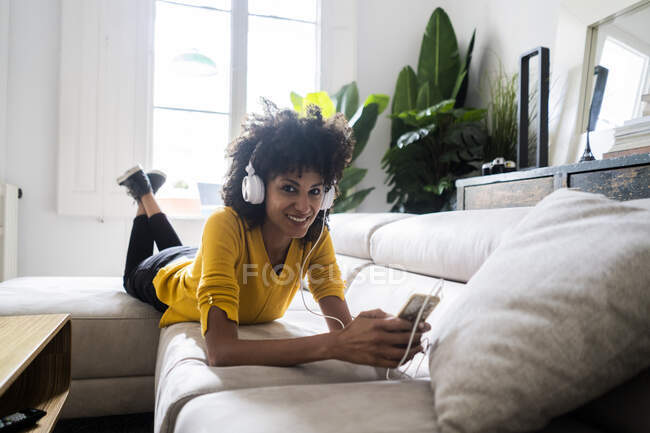 Porträt einer lächelnden Frau, die mit Handy und Kopfhörer auf dem Sofa liegt — Stockfoto