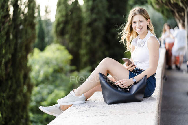 Ritratto di giovane donna sorridente con cuffie e smartphone seduta su un muro — Foto stock