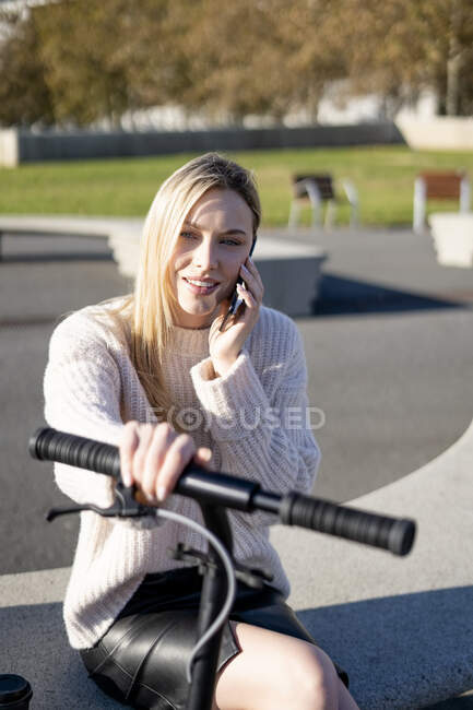 Portrait of young woman on the phone sitting on bench at sunlight with kick scooter — Stock Photo