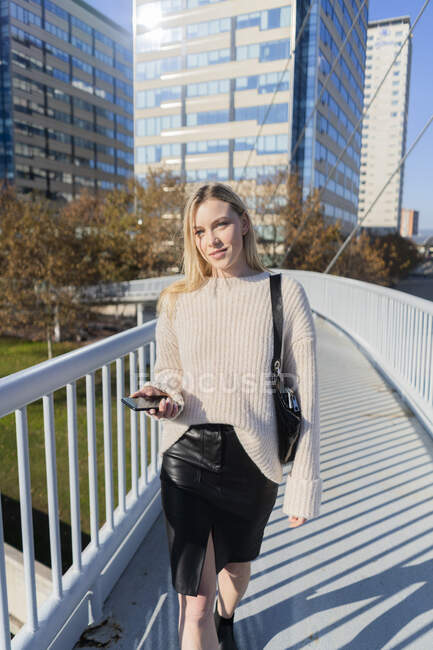 Ritratto di giovane donna bionda con smartphone che cammina sul ponte pedonale — Foto stock