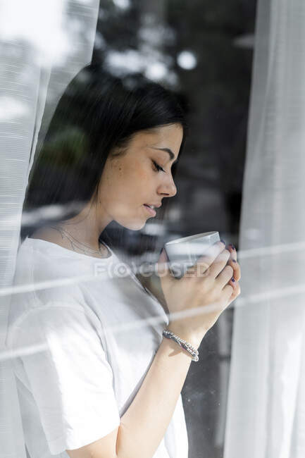 Mujer joven con taza de café detrás del cristal de la ventana - foto de stock