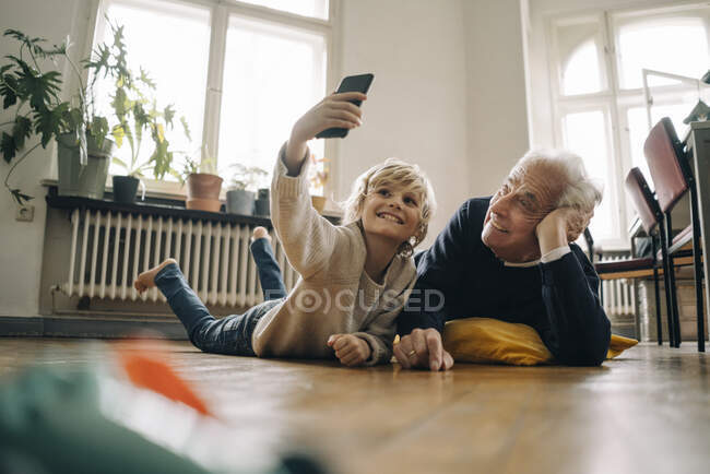 Grand-père et petit-fils allongés sur le sol à la maison prenant un selfie — Photo de stock
