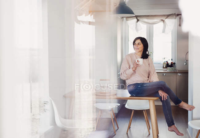Donna matura seduta sul tavolo della cucina, che beve caffè — Foto stock