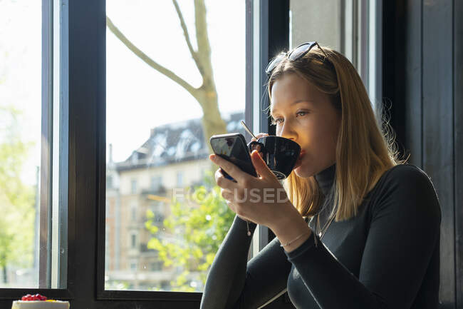 Молода жінка в кав'ярні п'є каву, дивлячись на смартфон. — стокове фото