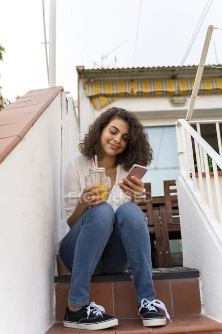 Giovane donna sorridente seduta sulle scale con cella e succo d'arancia — Foto stock