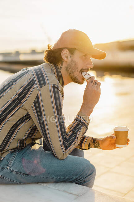 Tatuado homem com café para ir comer donut ao pôr-do-sol — Fotografia de Stock