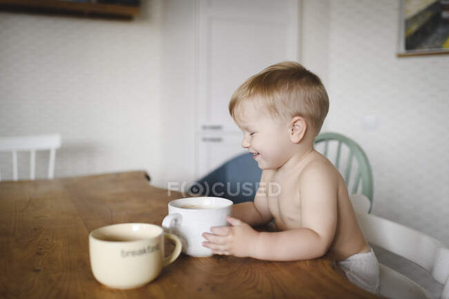 Lachender kleiner Junge sitzt am Küchentisch und hält Tasse — Stockfoto