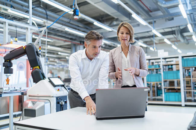 Homme d'affaires et femme d'affaires avec ordinateur portable parlant dans une salle d'usine moderne — Photo de stock