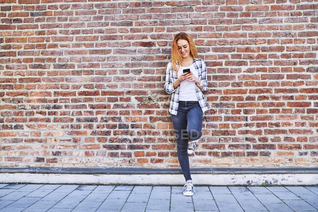 Giovane donna appoggiata al muro di mattoni in città durante l'utilizzo di smartphone — Foto stock