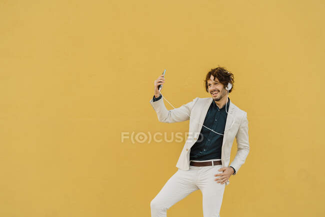 Uomo d'affari che balla davanti al muro giallo ascoltando musica con cuffie e smartphone — Foto stock