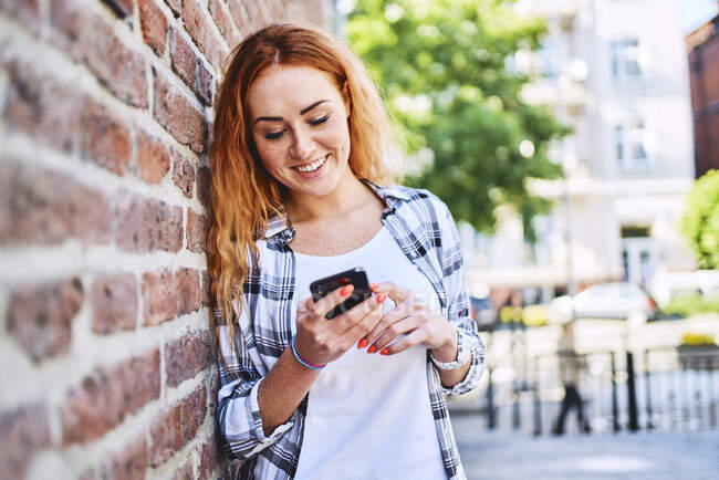 Young woman leaning against brick wall in the city while using smartphone — Stock Photo