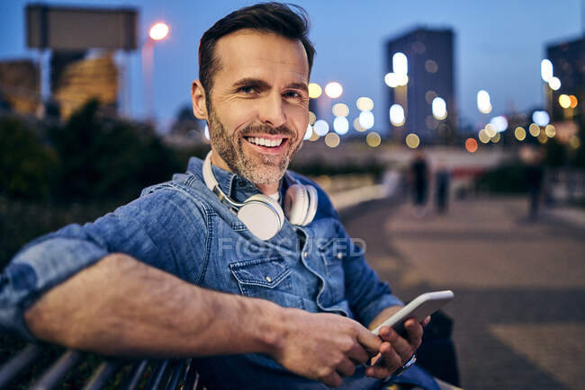 Retrato de un hombre sonriente usando su teléfono inteligente mientras está sentado en un banco por la noche - foto de stock