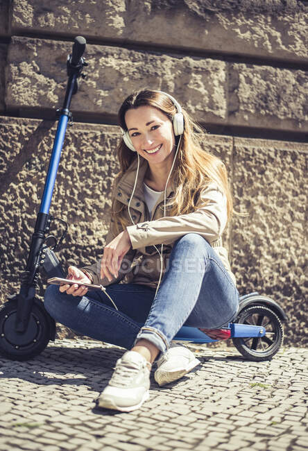 Ritratto di donna felice seduta su E-Scooter ad ascoltare musica con cuffie e smartphone — Foto stock